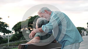 Middle age woman sitting on bench of park with old man hugging her from the back. Couple of retired people relaxing and enjoying