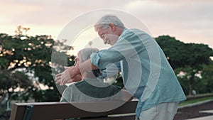 Middle age woman sitting on bench of park with old man hugging her from the back. Couple of retired people relaxing and enjoying