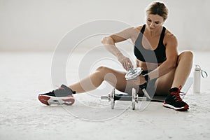 Middle age woman in sportswear puts disks on dumbbells sits on mat on floor