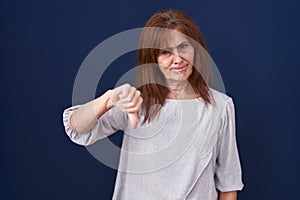 Middle age woman standing over blue background looking unhappy and angry showing rejection and negative with thumbs down gesture