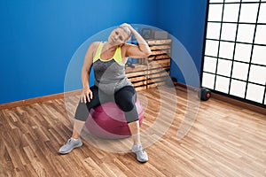 Middle age woman stretching neck sitting on fit ball at sport center