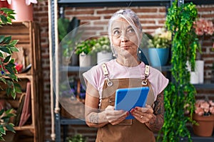 Middle age woman with tattoos working at florist shop with tablet smiling looking to the side and staring away thinking