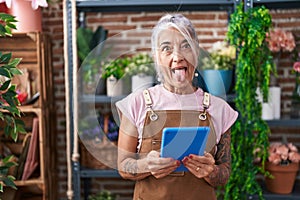 Middle age woman with tattoos working at florist shop with tablet sticking tongue out happy with funny expression