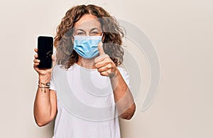 Middle age woman wearing coronavirus protection mask showing smartphone blank screen smiling happy and positive, thumb up doing