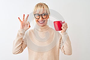 Middle age woman wearing glasses drinking red cup of coffee over isolated white background doing ok sign with fingers, excellent