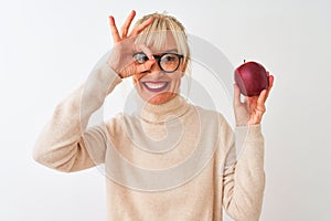Middle age woman wearing glasses holding apple standing over isolated white background with happy face smiling doing ok sign with