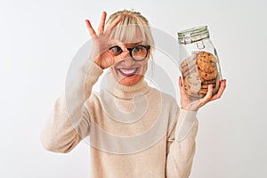 Middle age woman wearing glasses holding jar of cookies over isolated white background with happy face smiling doing ok sign with