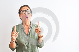 Middle age woman wearing green shirt and glasses standing over isolated white background amazed and surprised looking up and