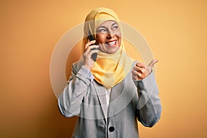 Middle age woman wearing traditional muslim hijab having conversation talking on smartphone pointing and showing with thumb up to