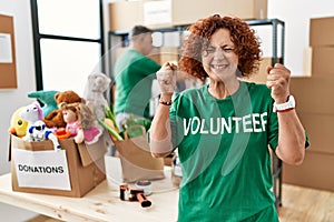 Middle age woman wearing volunteer t shirt at donations stand excited for success with arms raised and eyes closed celebrating