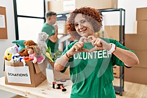 Middle age woman wearing volunteer t shirt at donations stand smiling in love doing heart symbol shape with hands