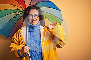 Middle age woman wearing yellow raincoat under colorful umbrella over isolated background with surprise face pointing finger to