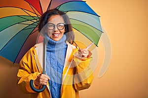 Middle age woman wearing yellow raincoat under colorful umbrella over isolated background very happy pointing with hand and finger