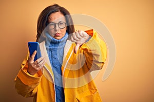 Middle age woman wearing yellow raincoat using smartphone over isolated yellow background with angry face, negative sign showing