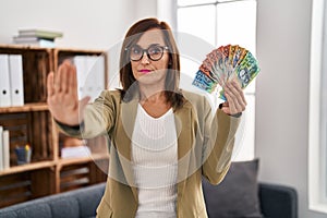 Middle age woman working at consultation office holding australian dollars with open hand doing stop sign with serious and