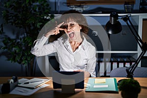 Middle age woman working at the office at night doing peace symbol with fingers over face, smiling cheerful showing victory