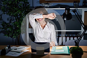 Middle age woman working at the office at night smiling cheerful playing peek a boo with hands showing face