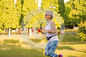Middle aged 40s or 50s happy and attractive woman with grey hair training at city park with green trees on sunrise doing running