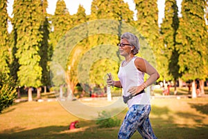 Middle aged 40s or 50s happy and attractive woman with grey hair training at city park with green trees on sunrise doing running