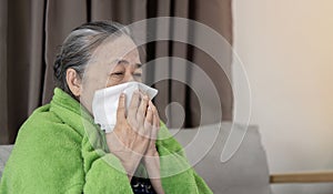 Middle-aged 60s sick frozen woman seated on sofa in living room covered with warm plaid sneezing holding paper napkin