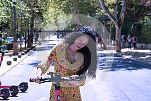 Middle-aged adult Hispanic woman with black curly hair, wearing a colorful outfit, leaning on an electric scooter. Concept scooter