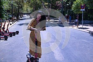 Middle-aged adult Hispanic woman with black curly hair, wearing a colorful outfit, leaning on an electric scooter. Concept scooter