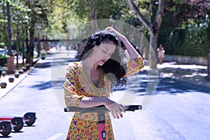 Middle-aged adult Hispanic woman with black curly hair, wearing a colorful outfit, leaning on an electric scooter. Concept scooter
