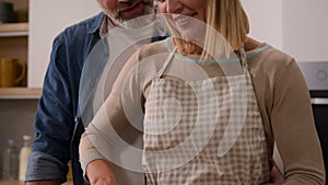 Middle-aged adult spouses cook vegetable salad in kitchen. Caring husband man hugging cuddling embracing loving wife