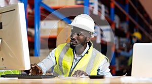 Middle aged African American warehouse worker working on personal computer and preparing a shipment in large warehouse