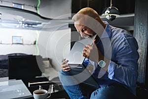 Middle aged angry Caucasian man, entrepreneur in casual clothes sitting on a table with a cup of coffee and holding a laptop in
