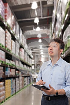 a middle-aged Asian businessman holding a tablet at a warehouse full of goods