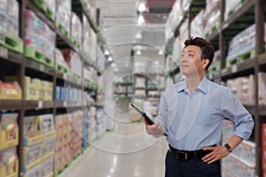 a middle-aged Asian businessman holding a tablet at a warehouse full of goods