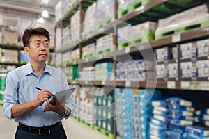 a middle-aged Asian businessman holding a tablet at a warehouse full of goods