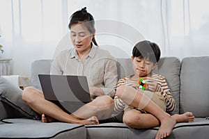 A middle-aged Asian single mother uses her laptop on the sofa in her home`s living room. The son ran and jumped onto the sofa and