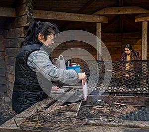 A middle-aged Asian woman cooking Souvlakia at a campsite fireplace.