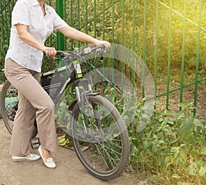 Middle-aged attractive slim woman in light trousers and shirt stands near the bike in the Park on a Sunny summer day, Cycling