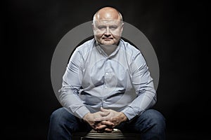 A middle-aged bald man sits on a chair. Black background