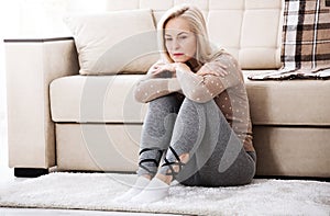 Middle aged barefoot woman sitting at the floor embracing her knees, near sofa at home, her head down, bored, troubled