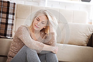 Middle aged barefoot woman sitting at the floor embracing her knees, near sofa at home, her head down, bored, troubled