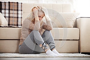 Middle aged barefoot woman sitting at the floor embracing her knees, near sofa at home, her head down, bored, troubled