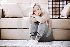 Middle aged barefoot woman sitting at the floor embracing her knees, near sofa at home, her head down, bored, troubled