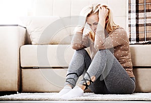 Middle aged barefoot woman sitting at the floor embracing her knees, near sofa at home, her head down, bored, troubled