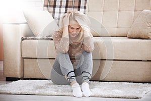 Middle aged barefoot woman sitting at the floor embracing her knees, near sofa at home, her head down, bored, troubled