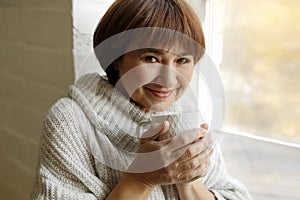 Middle aged beautiful mature woman in sweater drinking tea or coffee sitting on windowsill. Hygge concept