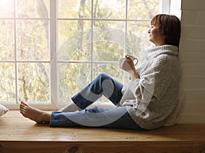 Middle aged beautiful mature woman in sweater drinking tea or coffee sitting on windowsill. Hygge concept