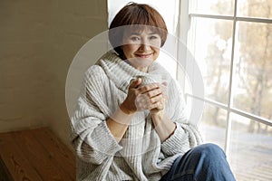 Middle aged beautiful mature woman in sweater drinking tea or coffee sitting on windowsill. Hygge concept