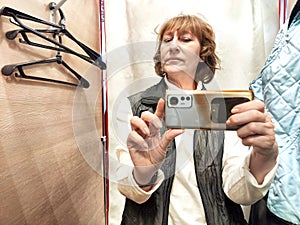 A middle-aged blonde woman in a cramped fitting room of a store with a bunch of clothes and hangers takes a selfie in photo