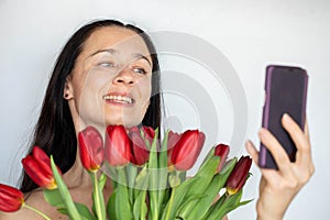 Middle aged brunette beautiful woman with a bouquet of red tulips taking selfie on phone. White background, copy space