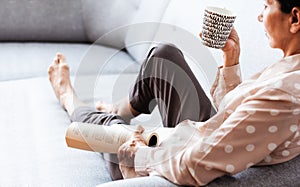 Middle-aged brunette woman on the gray sofa reading book with cup of coffee, soft focus, stay at home concept, cozy background