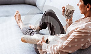 Middle-aged brunette woman on the gray sofa reading book with cup of coffee, soft focus, stay at home concept, cozy background